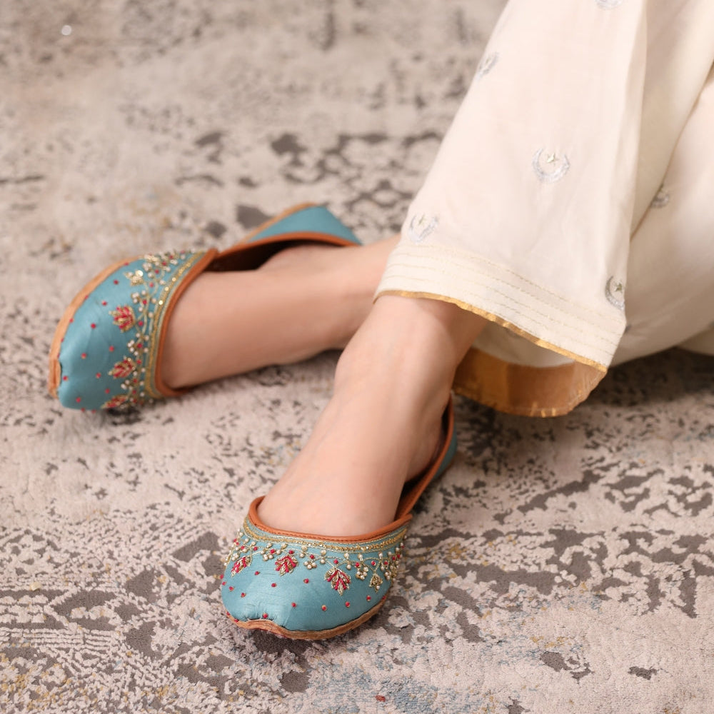 Blue khussas with floral patterns worn by a person on a textured floor.