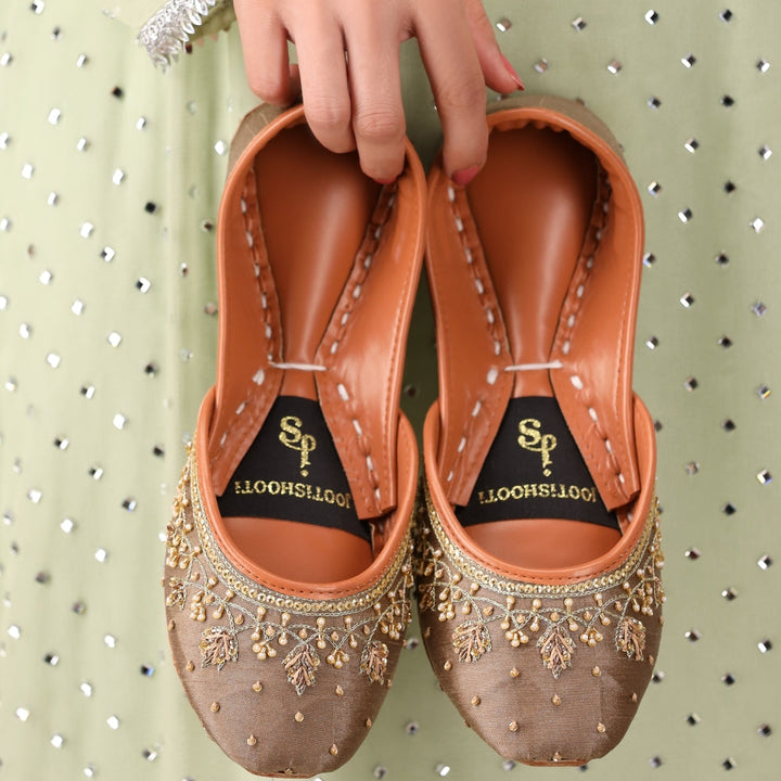 Pair of brown embellished shoes held by a hand against a light green background