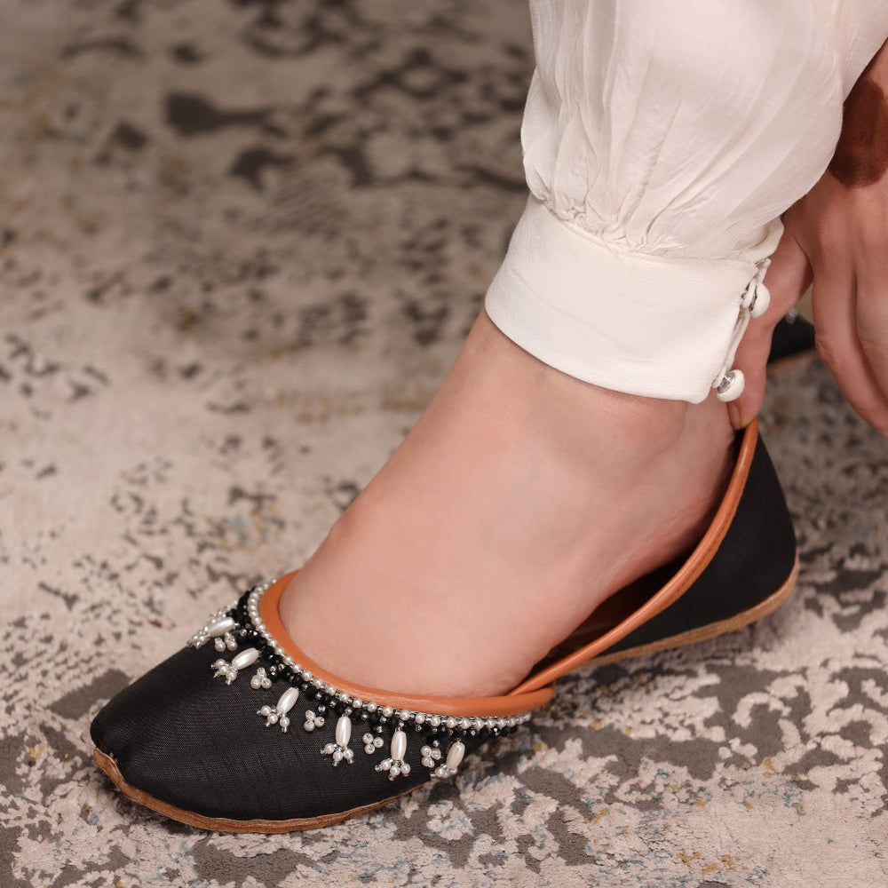 Black flat khussas with white embellishments worn by a person on a textured floor.