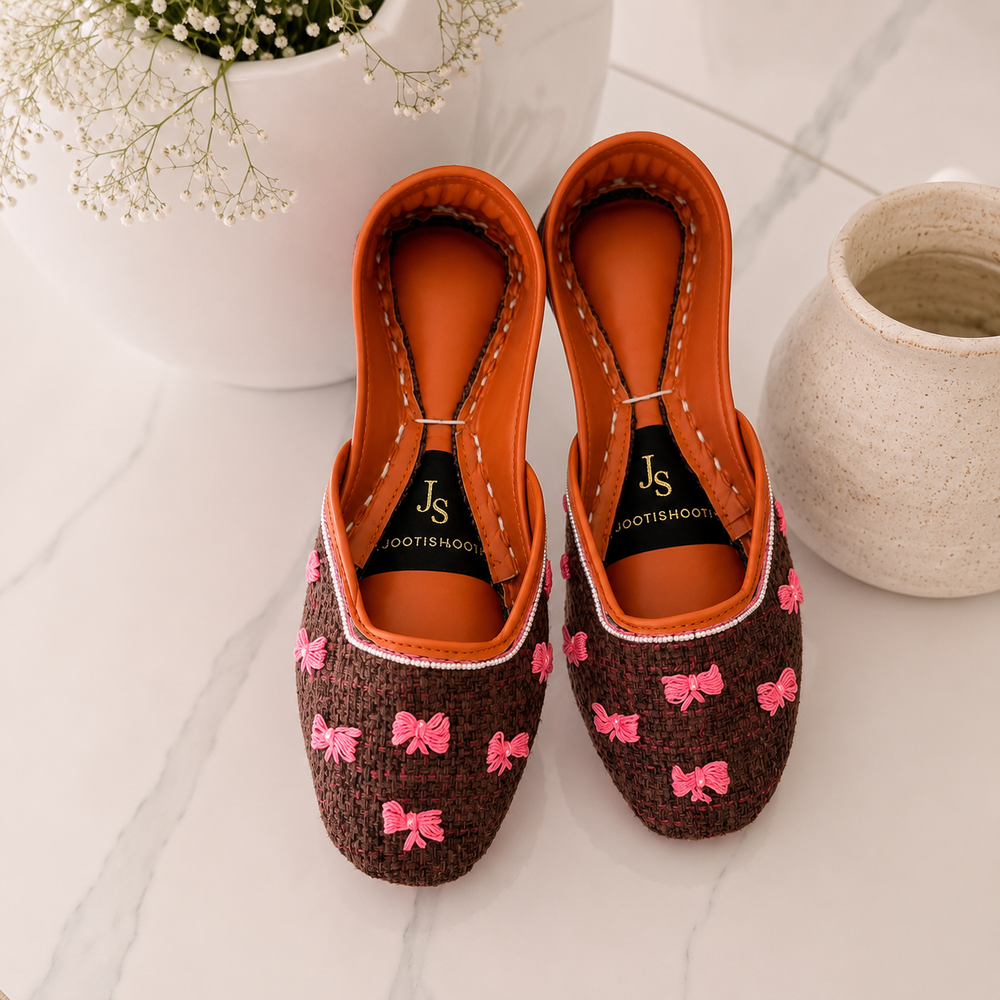 A pair of khussas with brown woven-texture uppers decorated with pink bow motifs, bright orange interiors, and black insoles branded 'Jootishooti', displayed on a light marble surface with decorative ceramic pots.