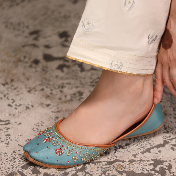 Blue embroidered khussas worn with light-colored pants on a textured floor.