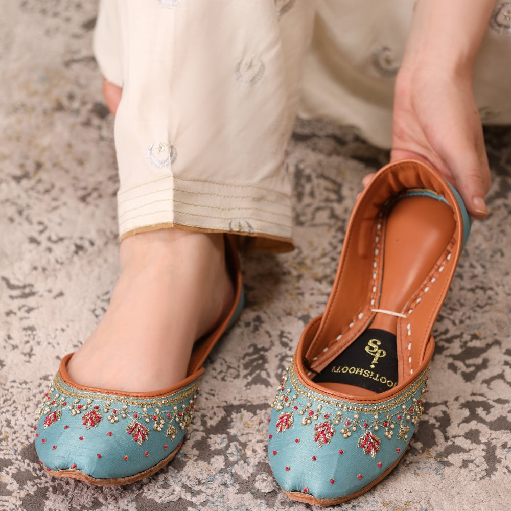Pair of traditional khussas with floral designs held by a person wearing beige pants.