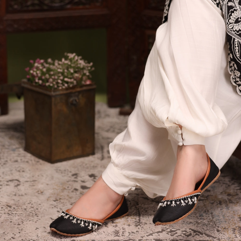 Person wearing black embellished khussas with white pants in a decorative setting.